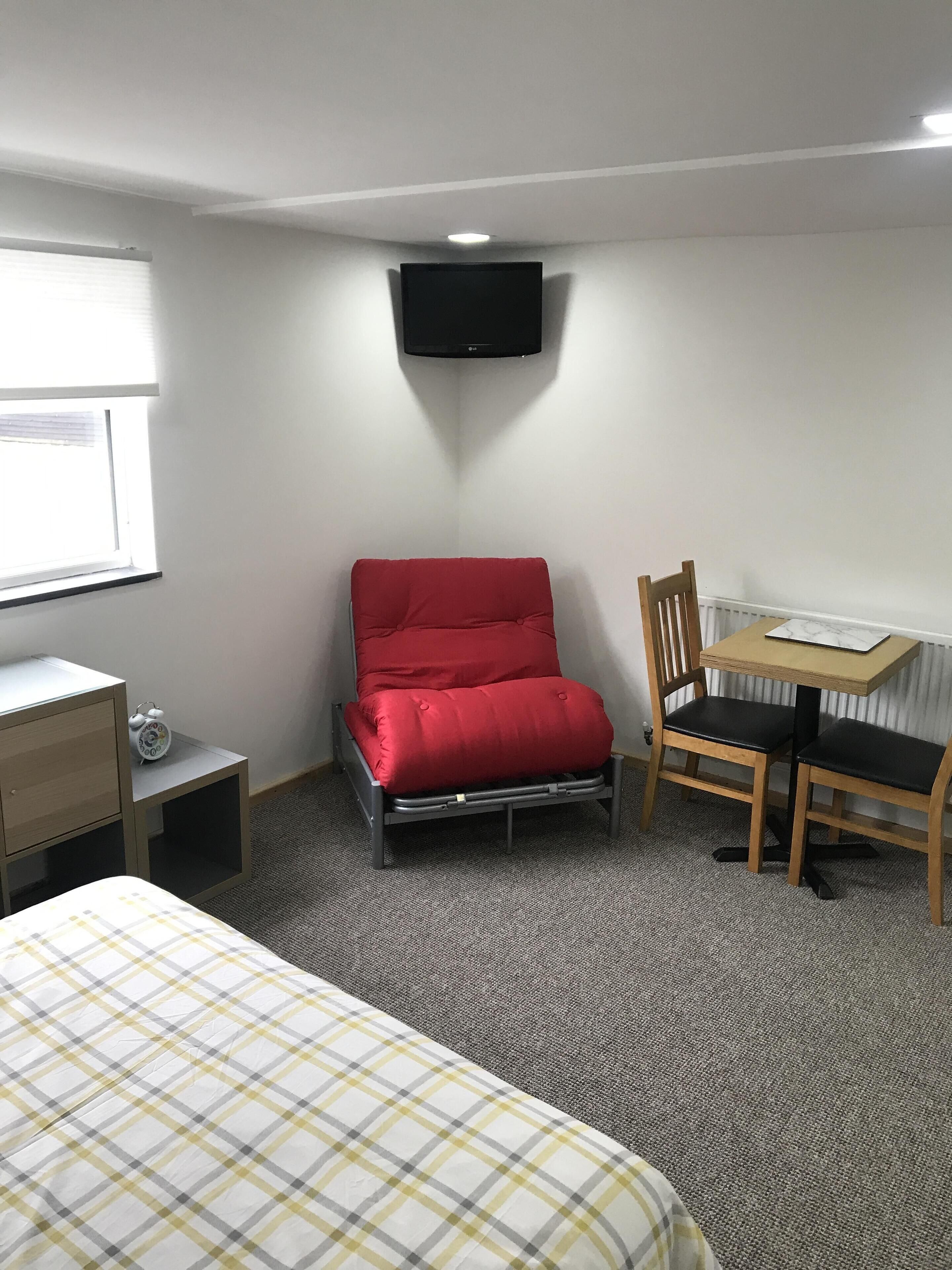 A bedroom with a bed, a desk with a chair, a red chair, a small table with a clock, and a TV on the wall.