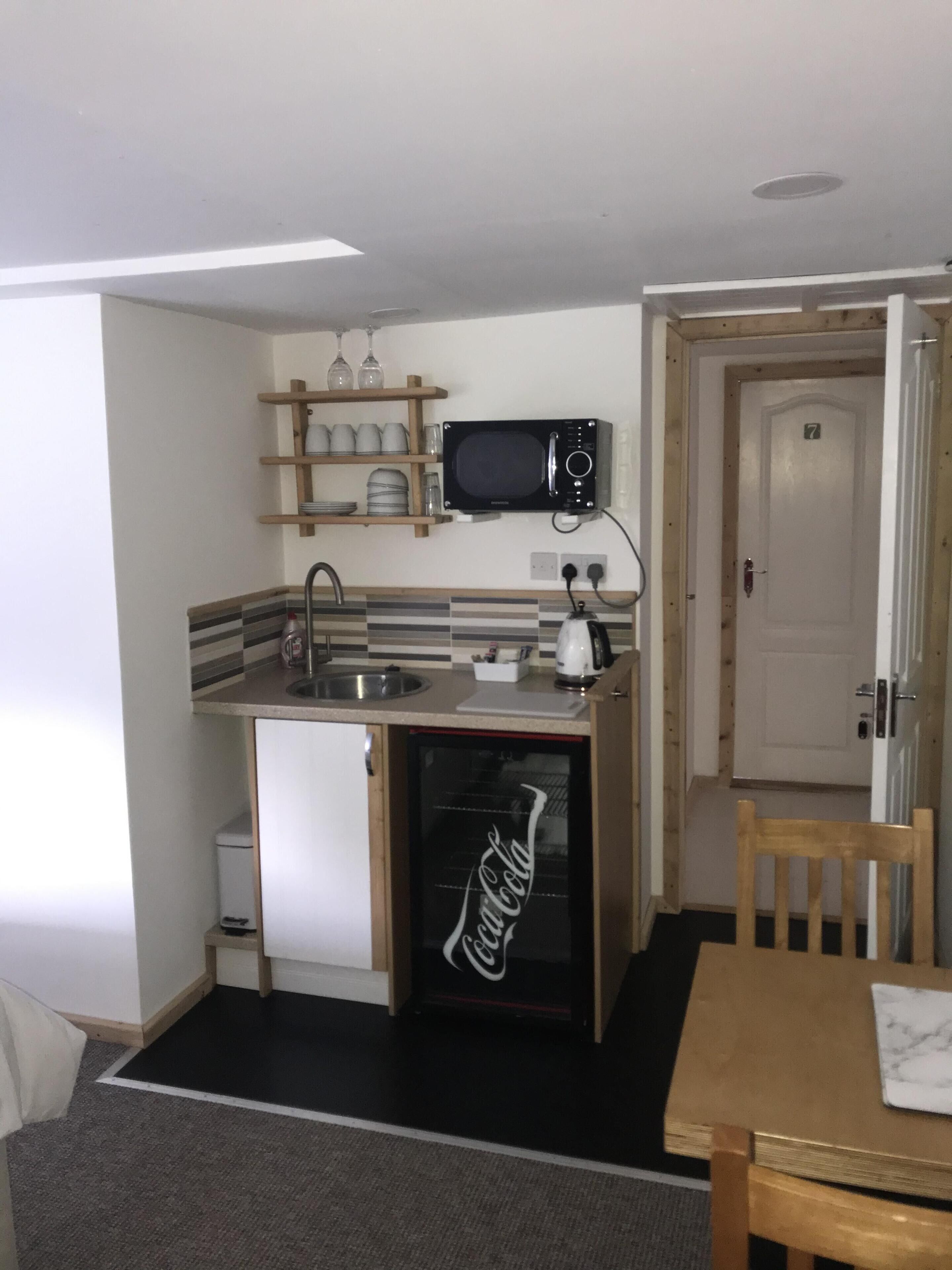Una cocina compacta con microondas, lavaplatos y refrigerador. Encima del microondas hay un estante de madera con platos y vasos. Se ve una puerta con el número 7 al fondo.