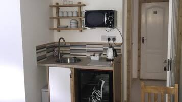 Una cocina compacta con microondas, lavaplatos y refrigerador. Encima del microondas hay un estante de madera con platos y vasos. Se ve una puerta con el número 7 al fondo.