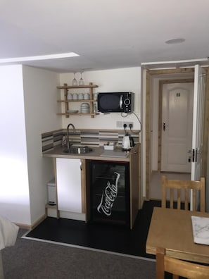A compact kitchen with a microwave, sink, and refrigerator. There is a wooden shelf above the microwave holding plates and glasses. A door with the number 7 is visible in the background. - Barton Gate Farm Guesthouse (Holsworthy)