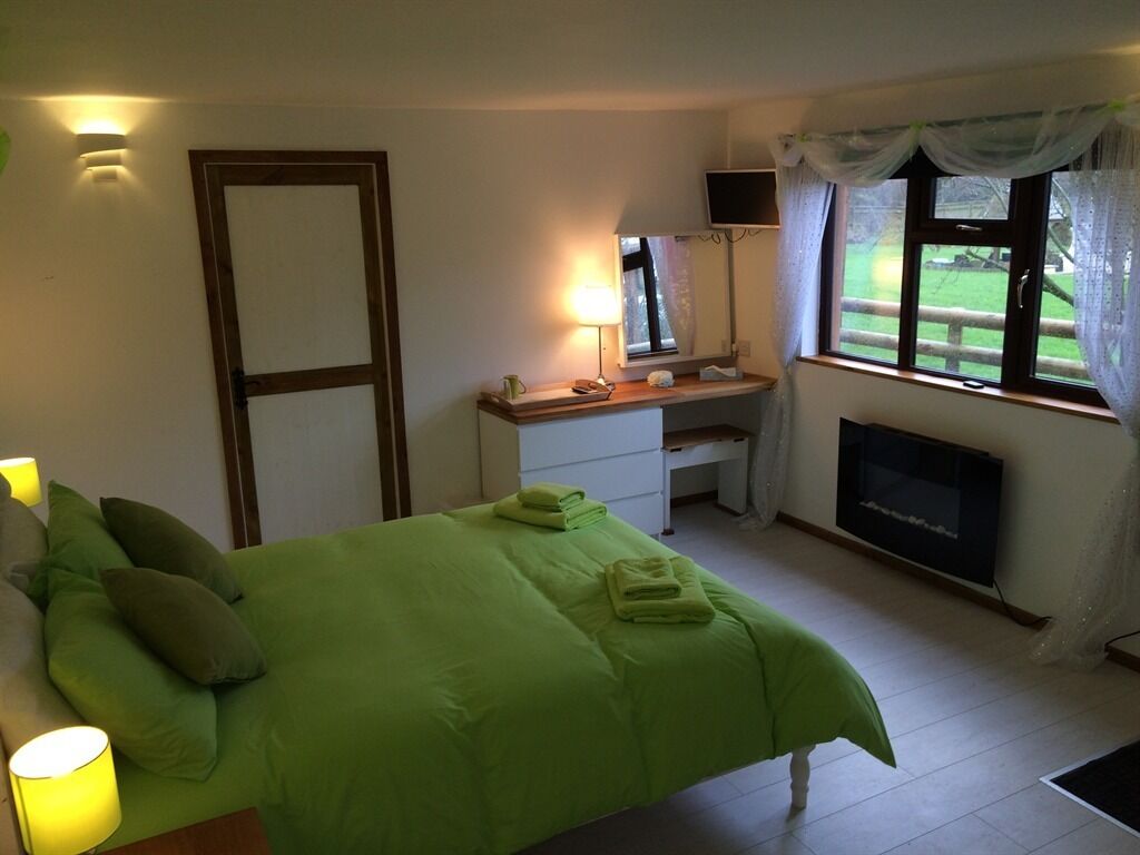 A bedroom with a green bed, a desk with a lamp, a TV, and a fireplace.