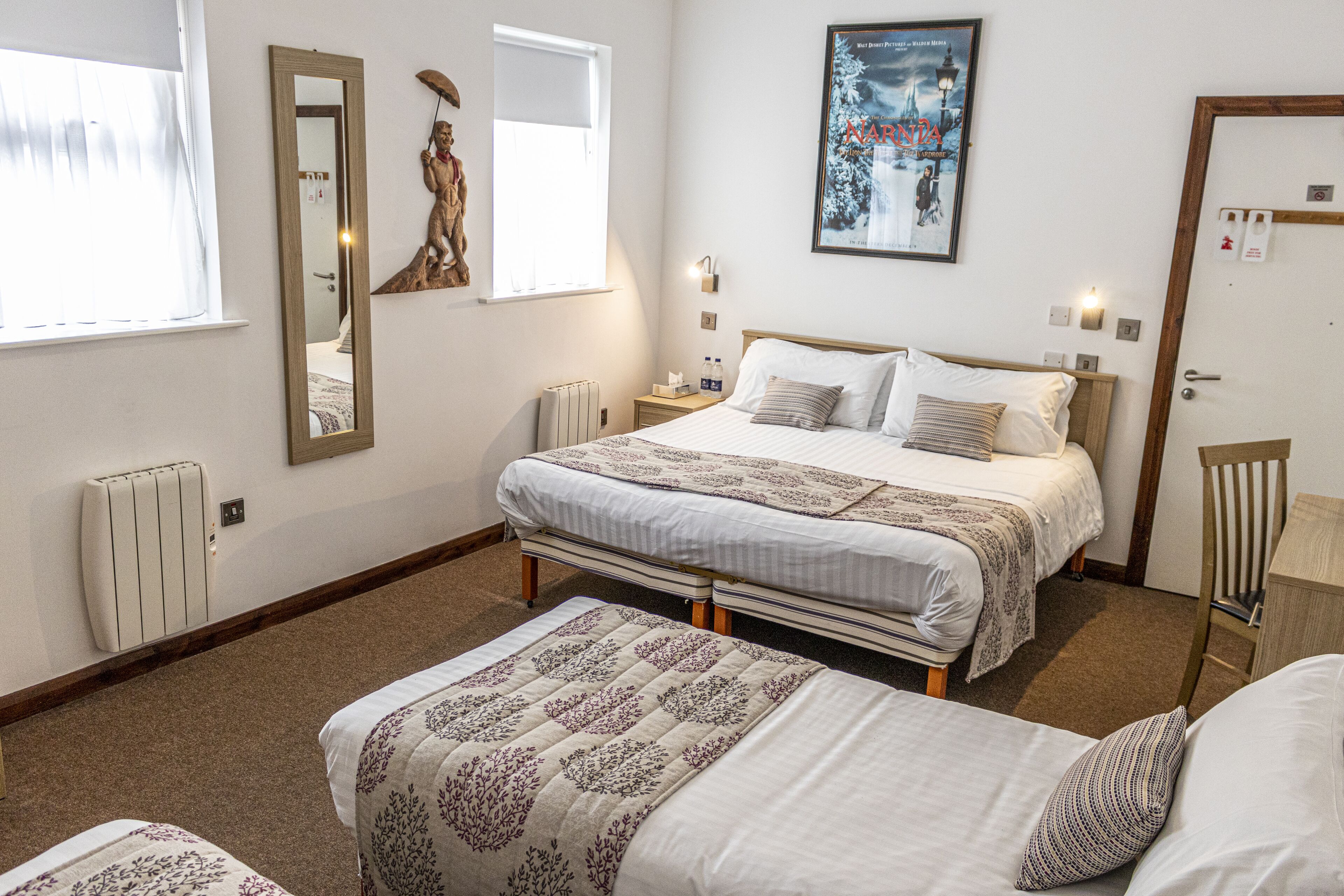 Habitación de hotel con una cama grande, otra más pequeña, una silla, un radiador, una ventana con cortinas, un espejo montado en la pared, un cuadro enmarco y una puerta con cerradura.
