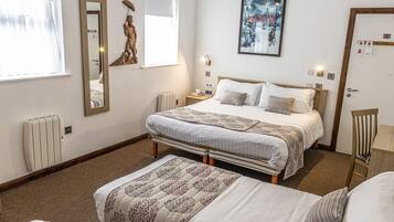 Habitación de hotel con una cama grande, otra más pequeña, una silla, un radiador, una ventana con cortinas, un espejo montado en la pared, un cuadro enmarco y una puerta con cerradura.