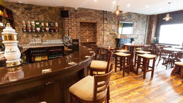 A bar area with a stone wall, wooden bar counter, and multiple stools. - The Rostrevor Inn (Newry)