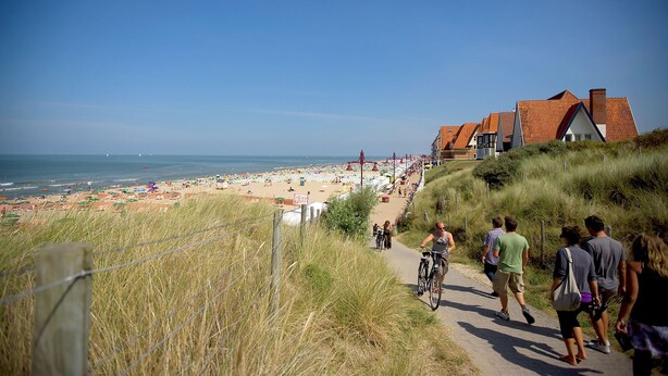 A coastal path leading to a beach with people walking and cycling, and houses with red roofs in the background. - Holiday Suites De Haan (De Haan)