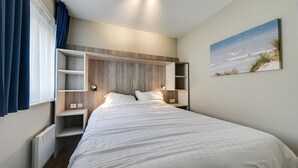 A neatly arranged bedroom with a bed, built-in shelving, bedside tables, and a painting of a beach scene. - Holiday Suites De Haan (De Haan)