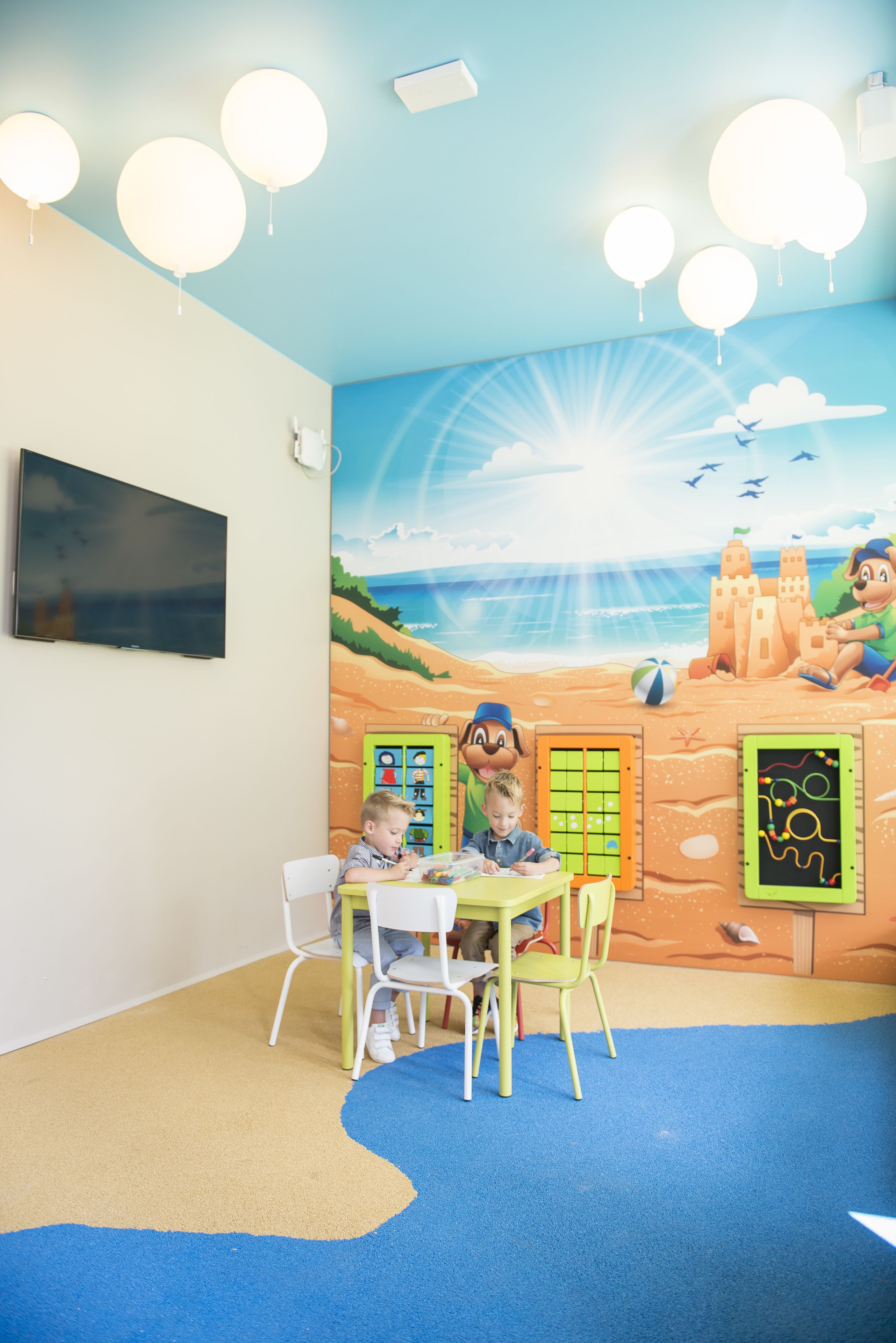 Uma sala de jogos infantil com um mural de praia, uma TV de tela plana e uma mesa com cadeiras.