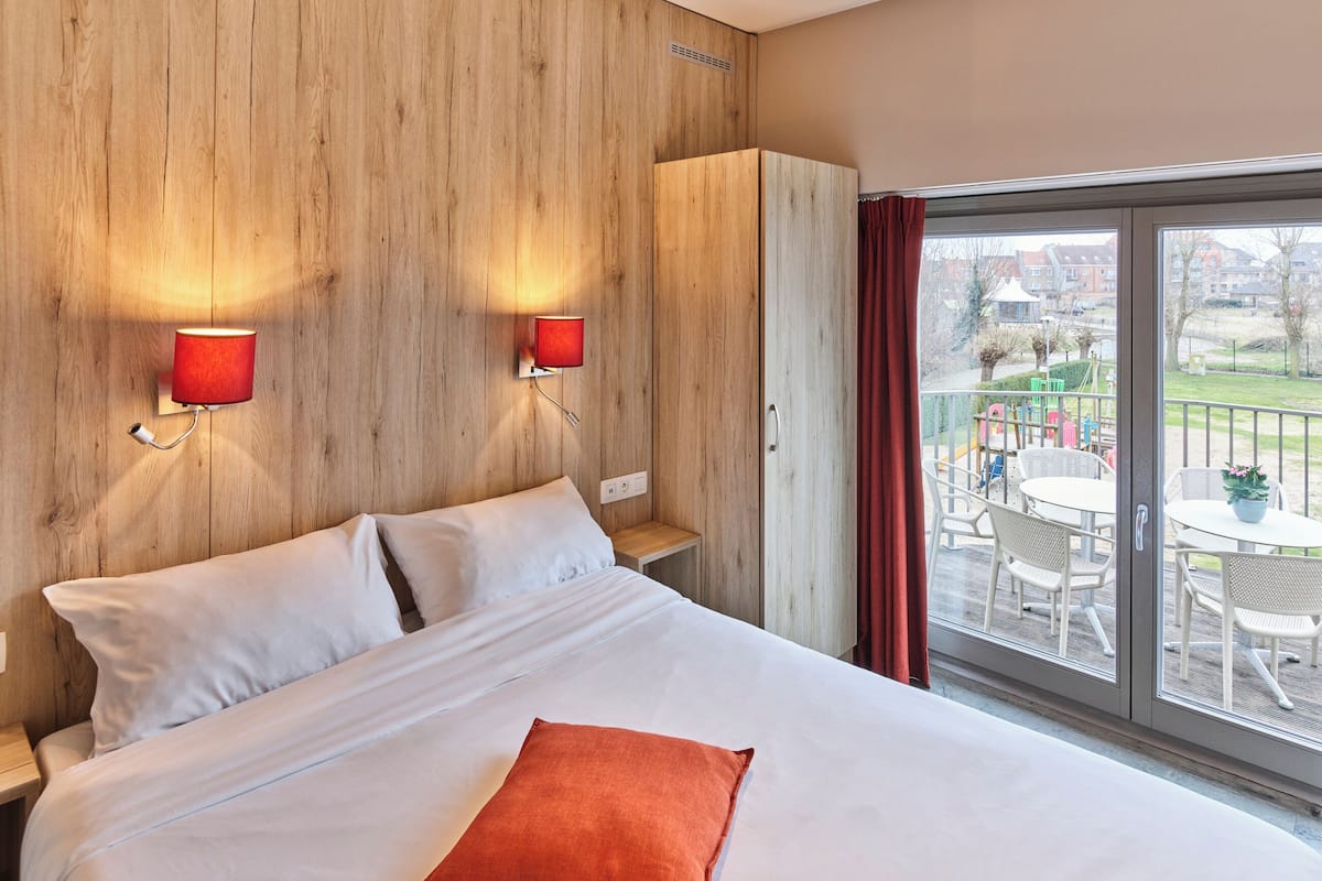 Une chambre d’hôtel avec un lit, un oreiller rouge, deux lampes fixées au mur et une vue sur un balcon aménagé avec du mobilier d’extérieur.