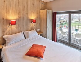 Habitación de hotel con una cama, un almohadón rojo, dos lámparas de pared y vistas a un balcón con mobiliario exterior.