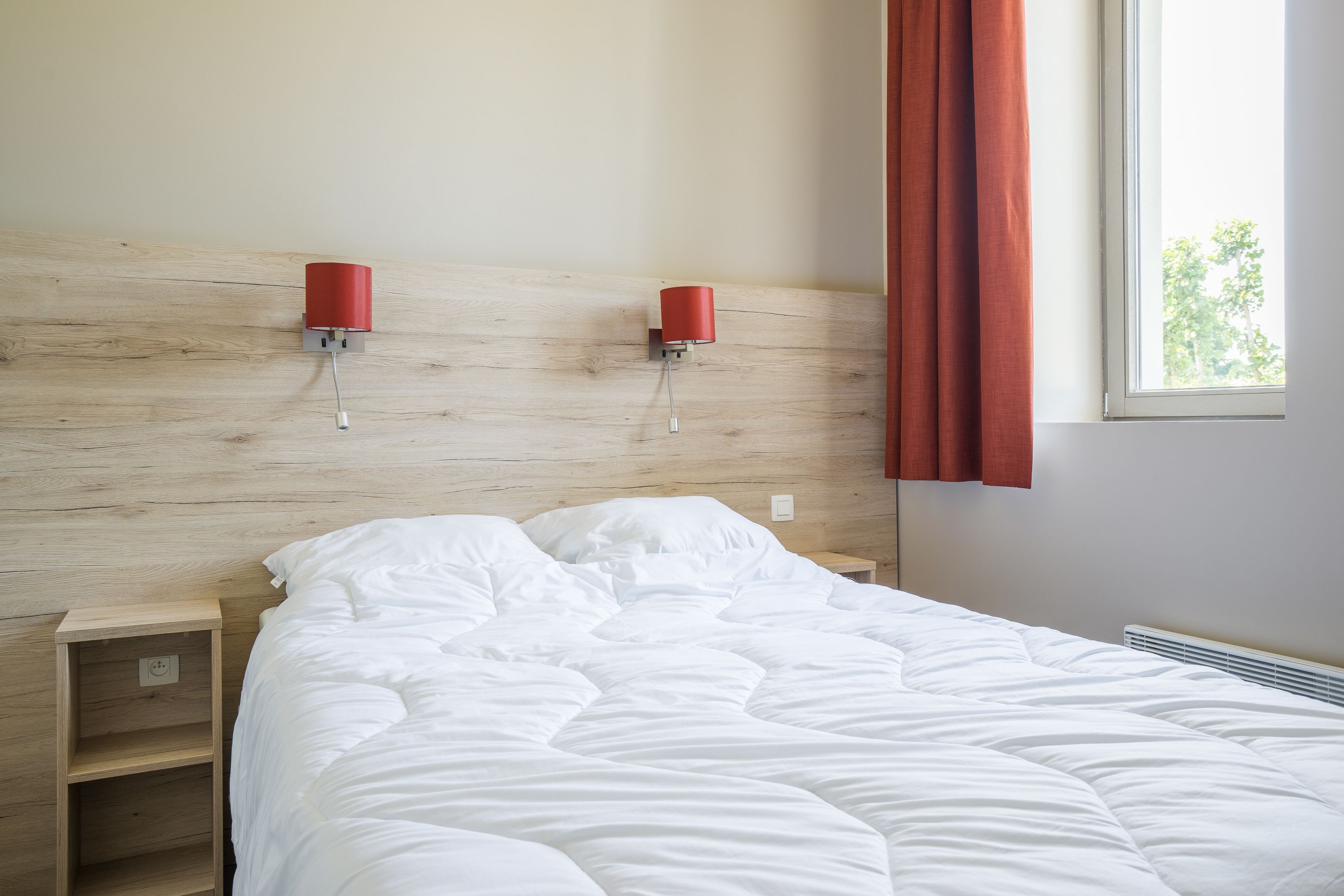 Una cama bien hecha con ropa blanca, un cabecero de madera y dos lámparas rojas fijadas a la pared.