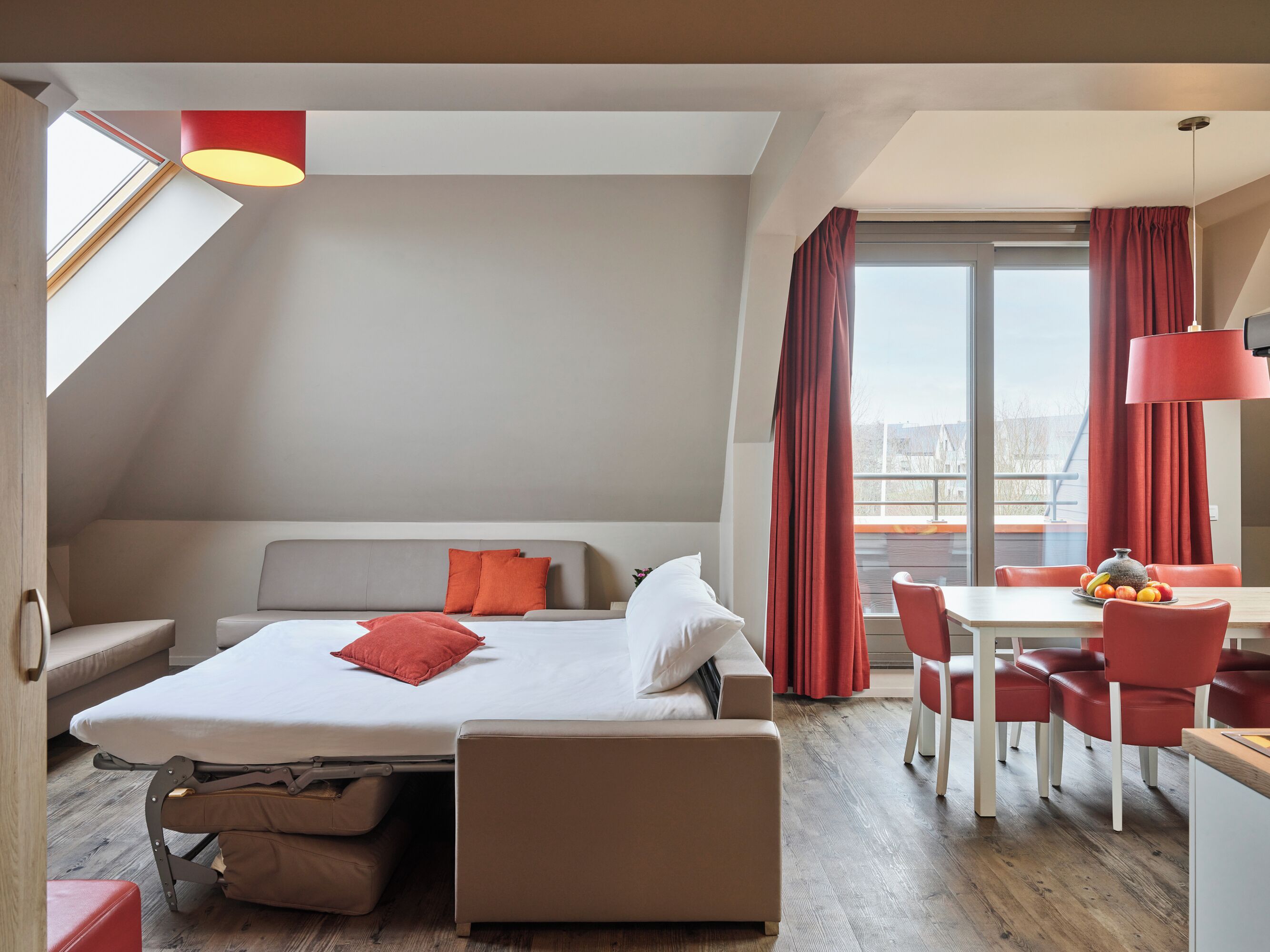 A modern hotel room with a loft bed, a sofa, a dining table with chairs, and a view of the city.