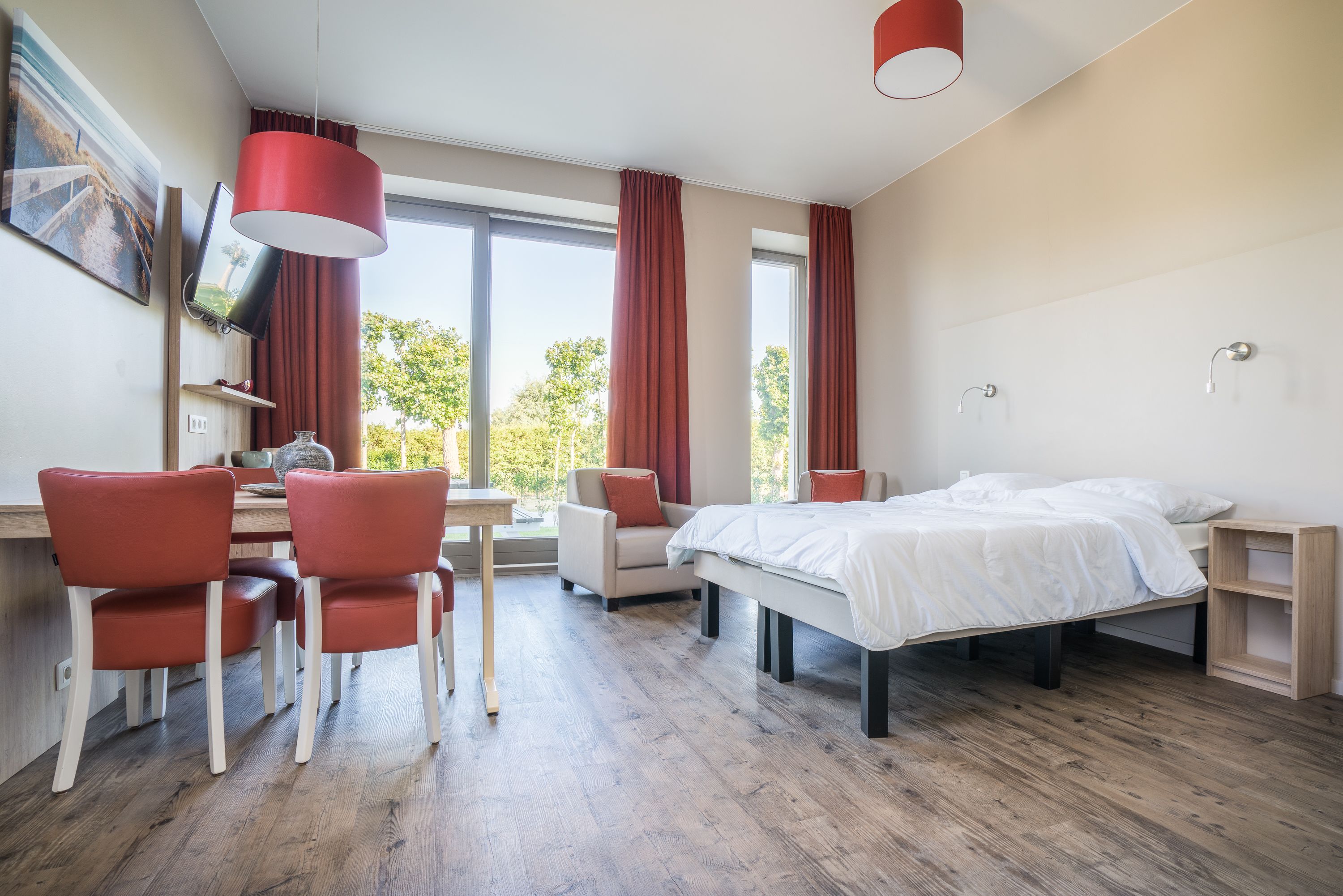 Moderni hotellihuone, jossa on puulattia, sänky, työpöytä tuoleineen, televisio ja suuret ikkunat, joissa on verhot.