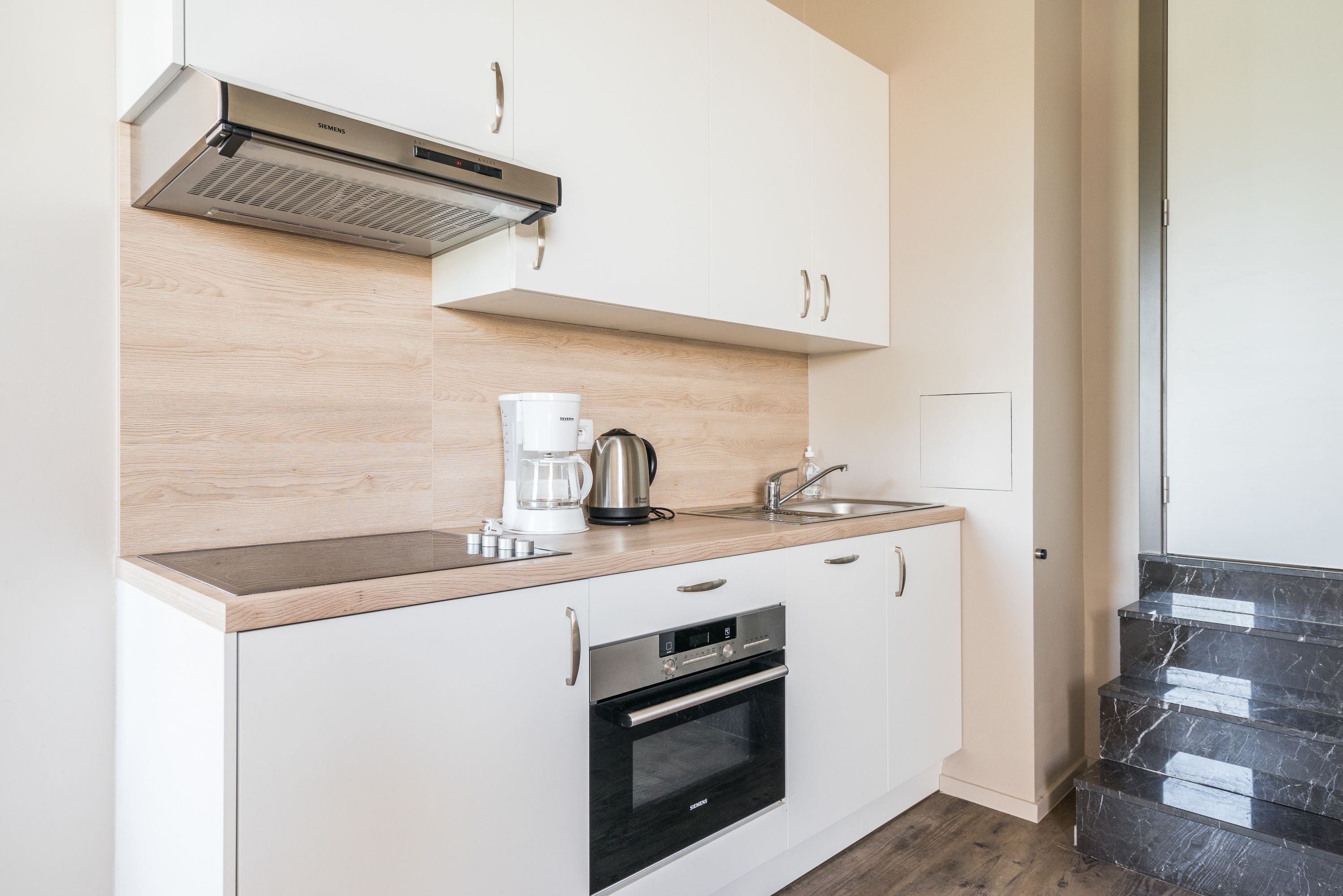 A modern kitchen with white cabinets, a stainless steel range hood, a coffee maker, and a built-in oven.