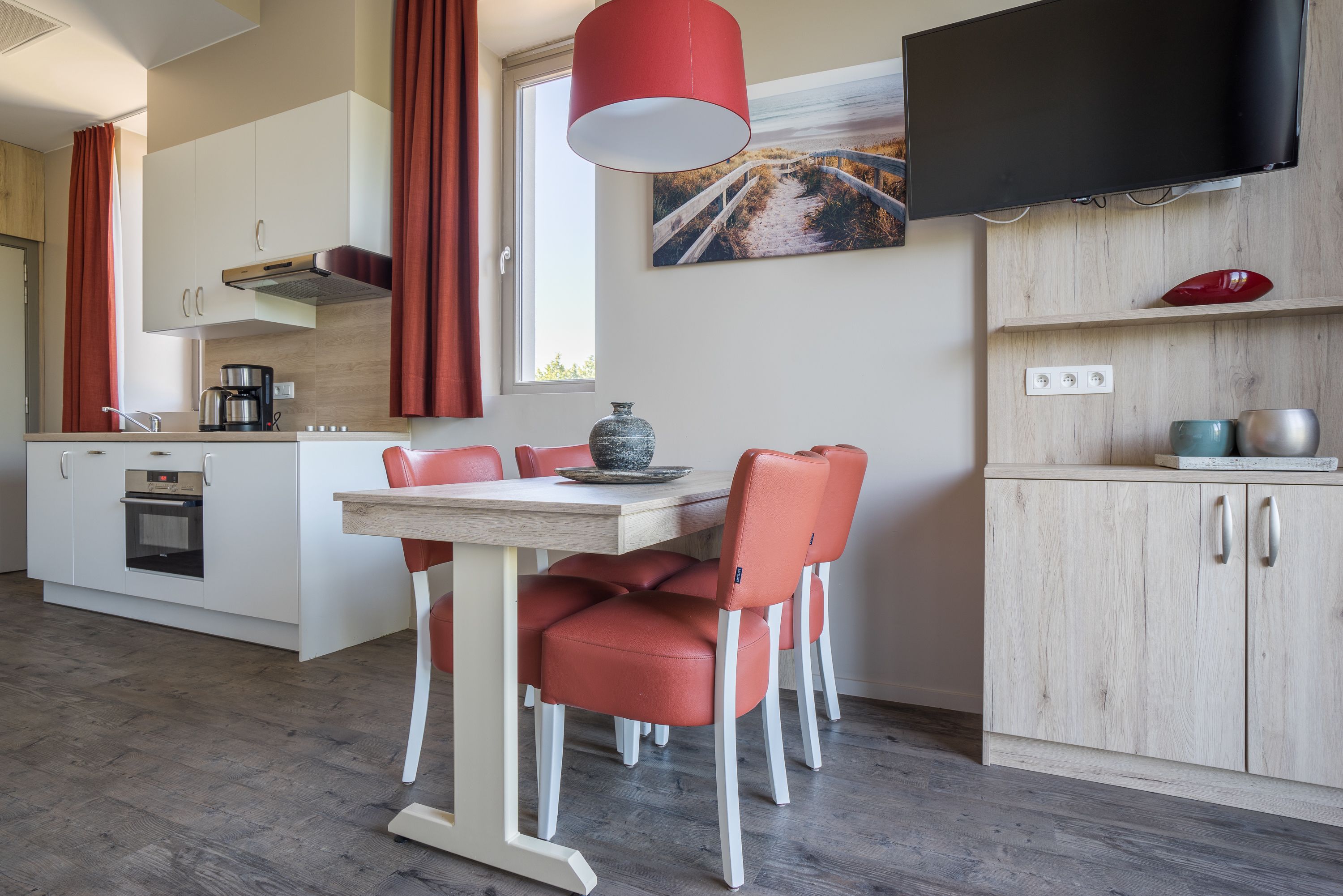 Une cuisine moderne avec des meubles blancs, une table à manger avec des chaises rouges, une machine à café et un téléviseur fixé au-dessus des meubles.