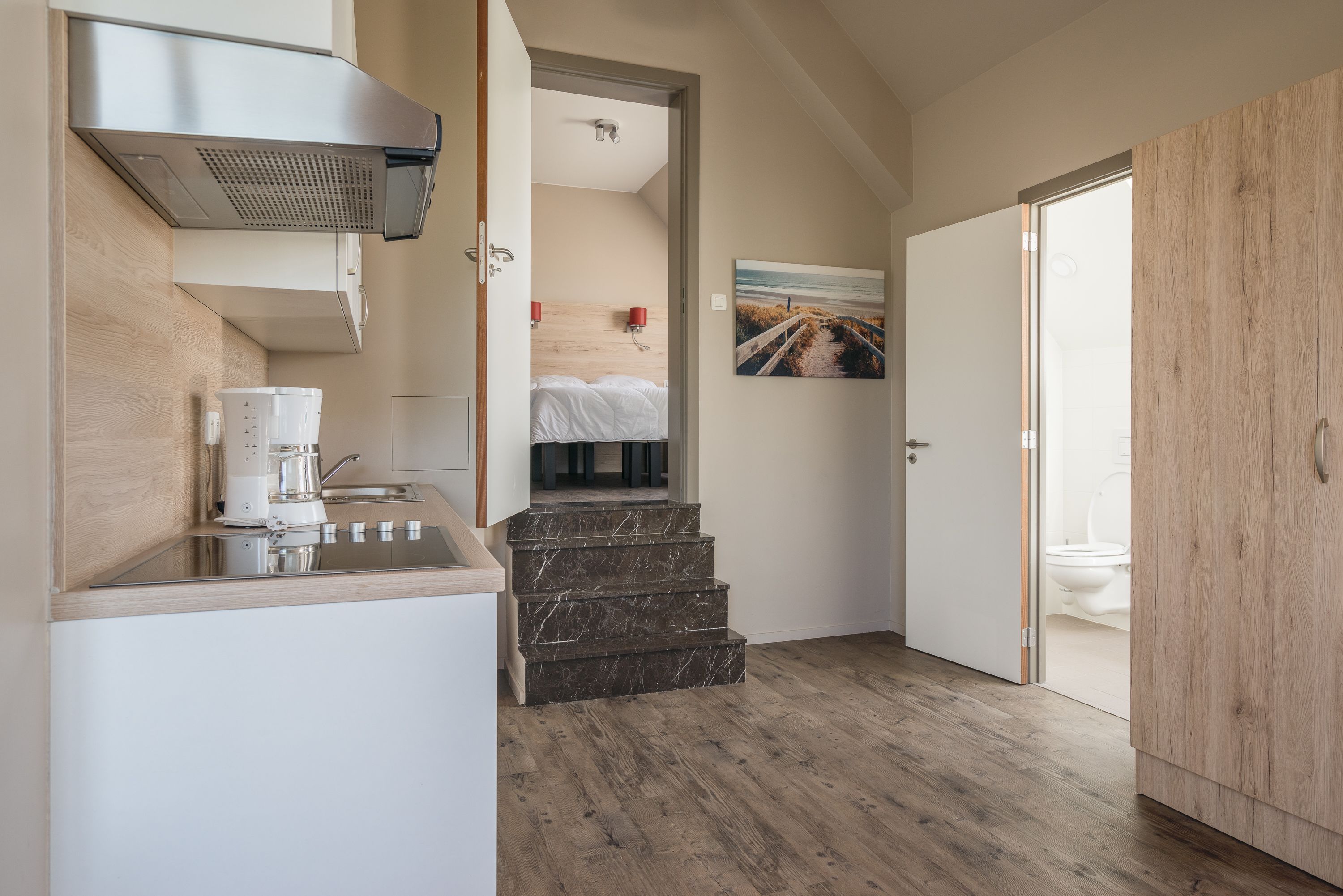 Een kleine keuken met een koffiezetapparaat, een fornuis en een spoelbak. Er zijn houten treden die leiden naar een slaapkamer met een bed en een schilderij van een strand.