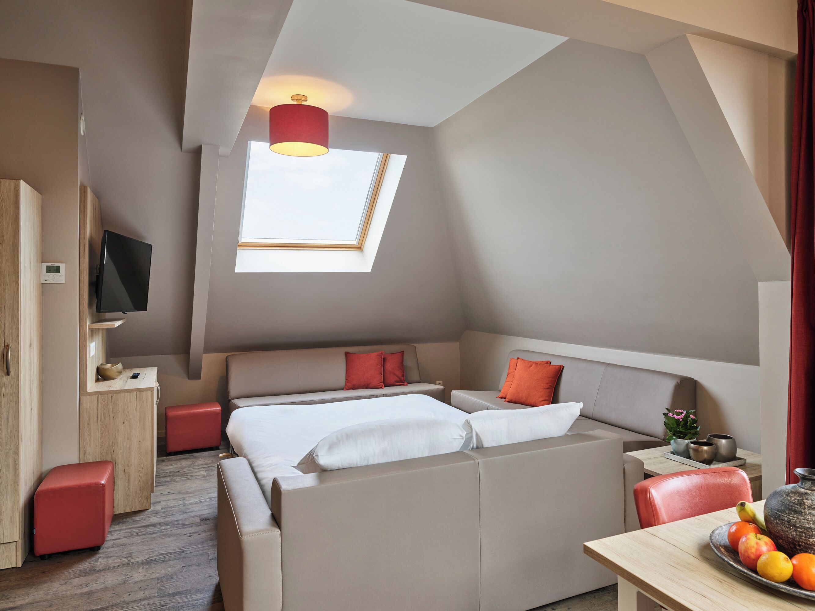 A modern loft bedroom with a sloped ceiling, a skylight, a bed with white bedding, a red accent pillow, a red ottoman, a wooden nightstand, a red chair, and a table with fruit and a vase.