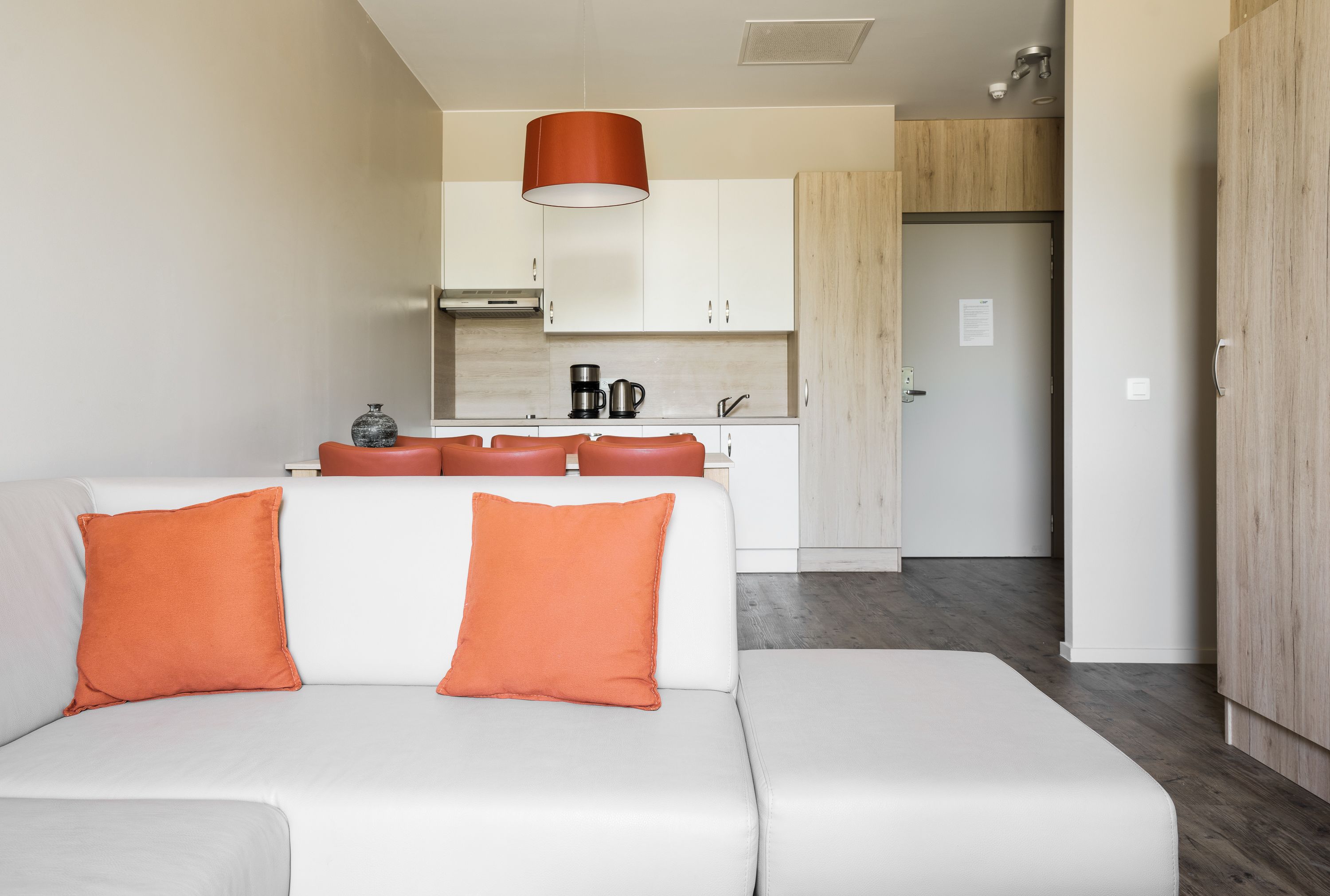 Una sala de estar moderna con un sofá blanco y cojines naranjas, una zona de cocina con armarios blancos y una cafetera, y una puerta de madera.