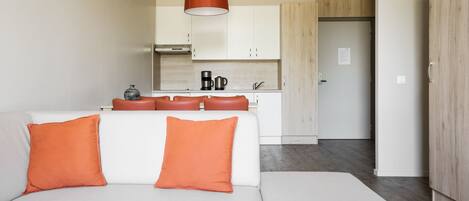 A modern living room with a white sofa and orange cushions, a kitchen area with white cabinets and a coffee machine, and a wooden door.