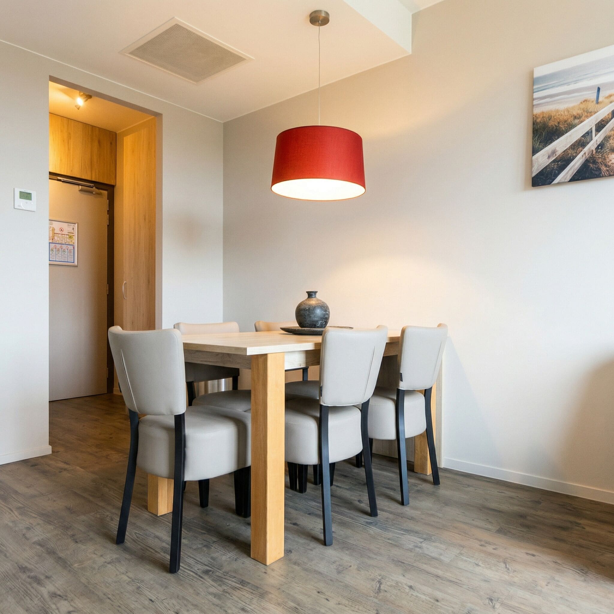 Un coin repas avec une table et des chaises en bois, un luminaire suspendu rouge et un tableau au mur.