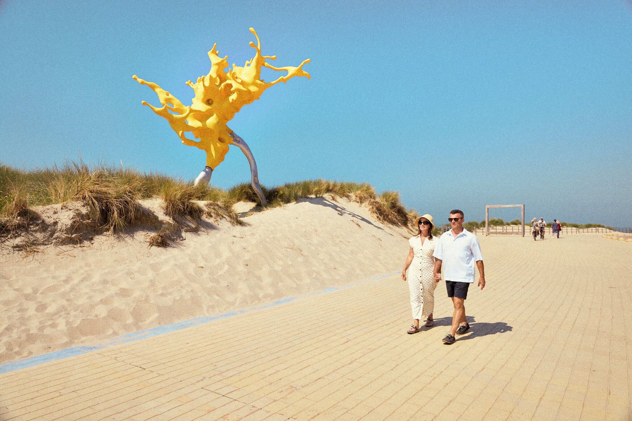 Una pareja pasea sobre una playa arenosa, con una llamativa escultura amarilla al fondo.