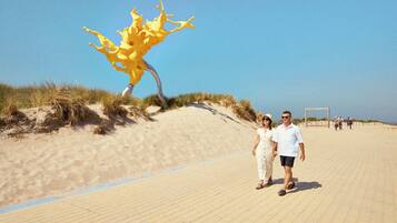 Ein Paar spaziert am sandigen Strand, im Hintergrund eine markante, gelbe Skulptur.
