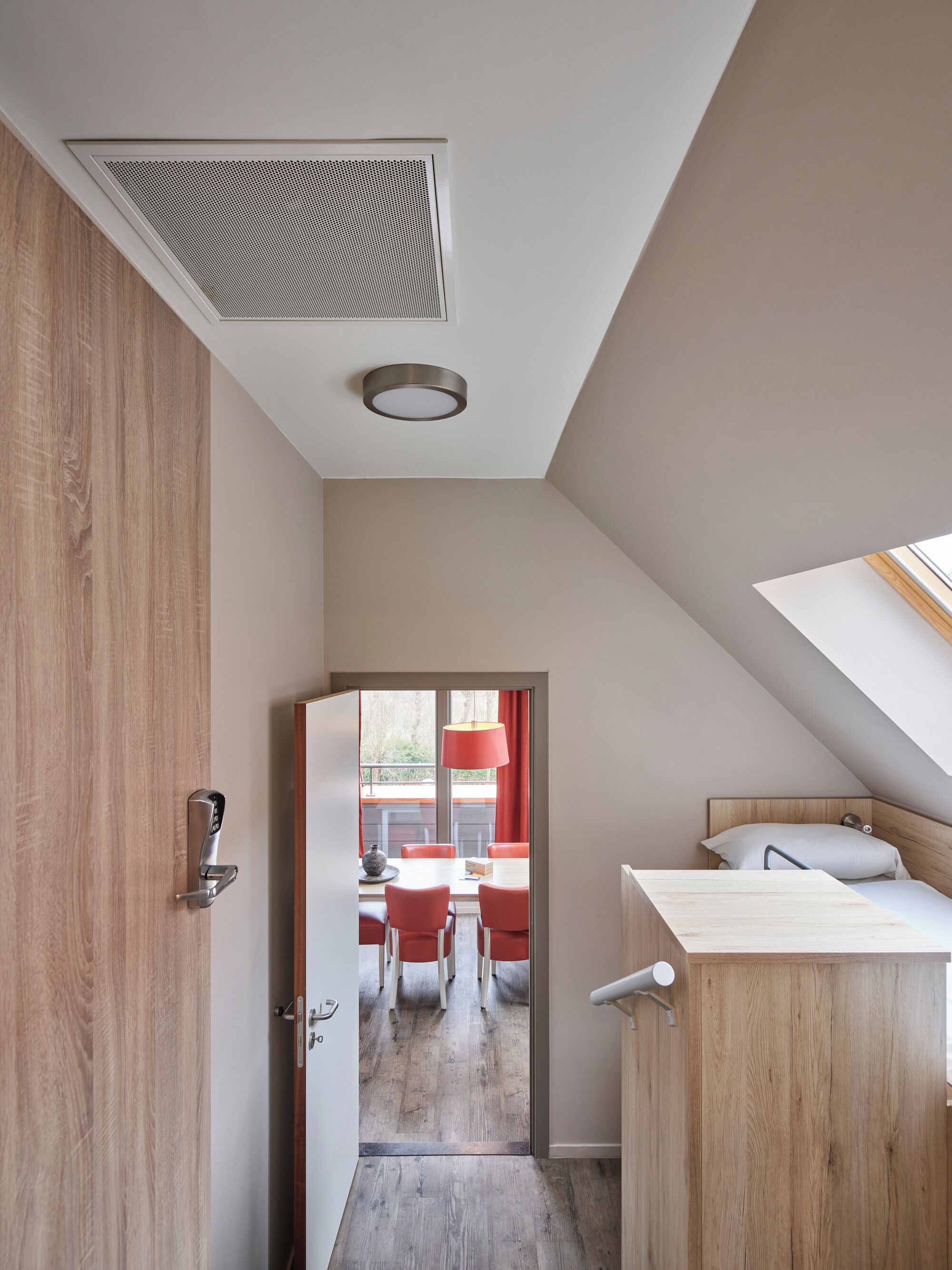 Una habitación moderna y compacta con puerta de madera, una pequeña kitchenette con ventana y un comedor con sillas rojas y una mesa.