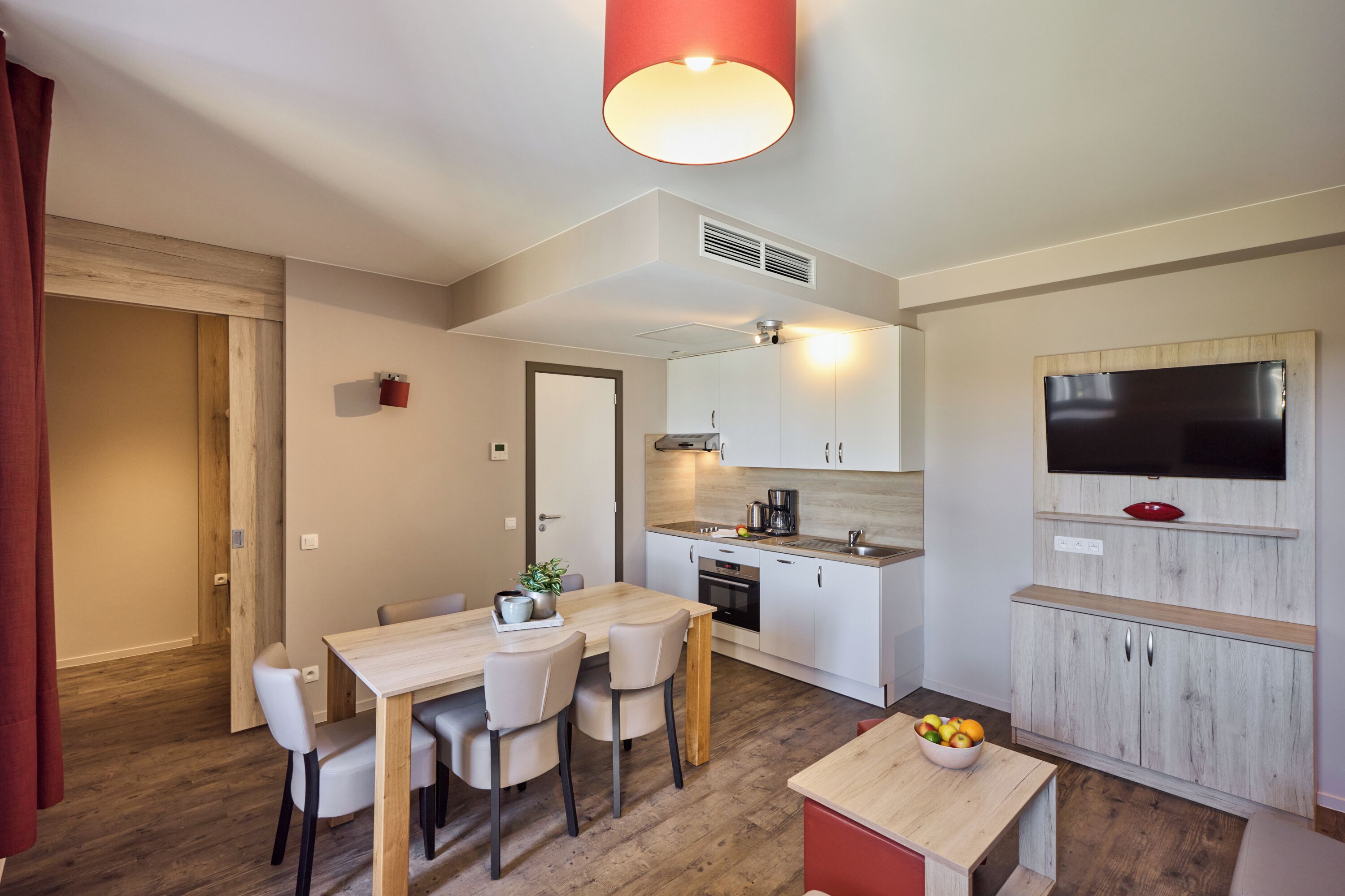 A modern kitchen with a dining area, wooden flooring, and a TV mounted on the wall.