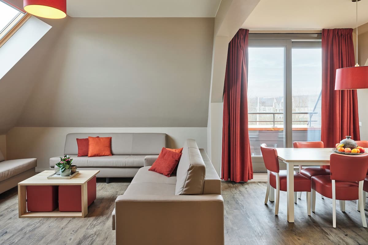 Un salon moderne avec un canapé gris, des coussins rouges, une table basse en bois et un coin repas comprenant une table blanche et des chaises rouges.