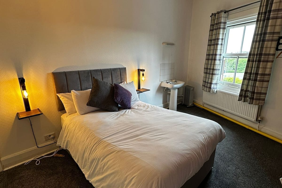 A bedroom with a bed, bedside tables, a nightstand, a window with curtains, and a radiator.