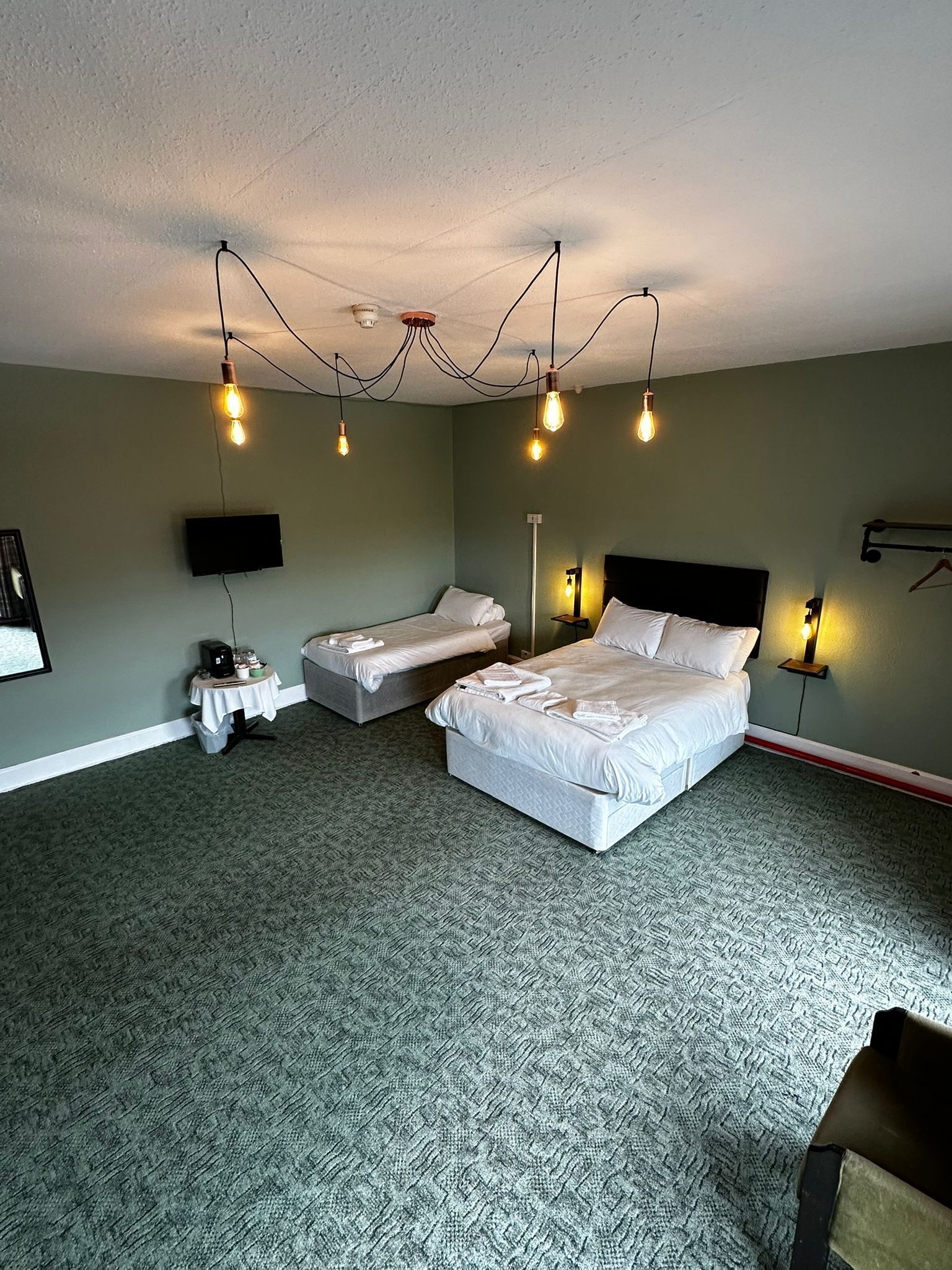 A hotel room with a large bed, two smaller beds, a flat-screen TV, a coffee machine, and a table with a tray of items.