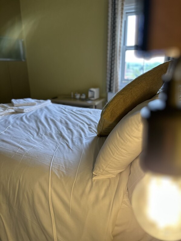 Una cama bien tendida con sábanas blancas y un cojín marrón, una mesita de noche con una lámpara, y una ventana con cortinas.