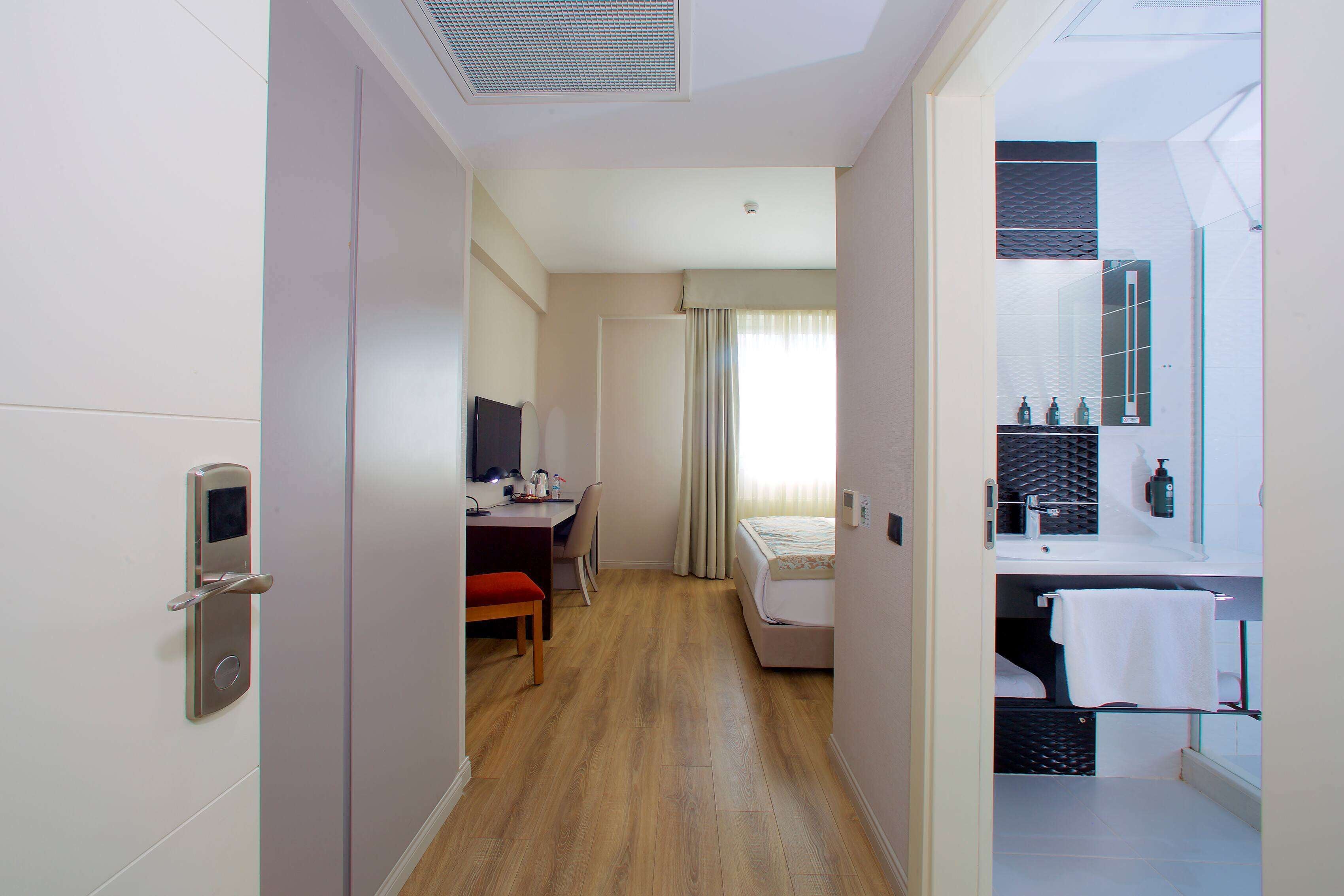 A hotel room with a wooden floor, a desk with a chair, a bed, a bathroom with a mirror and towel rack.