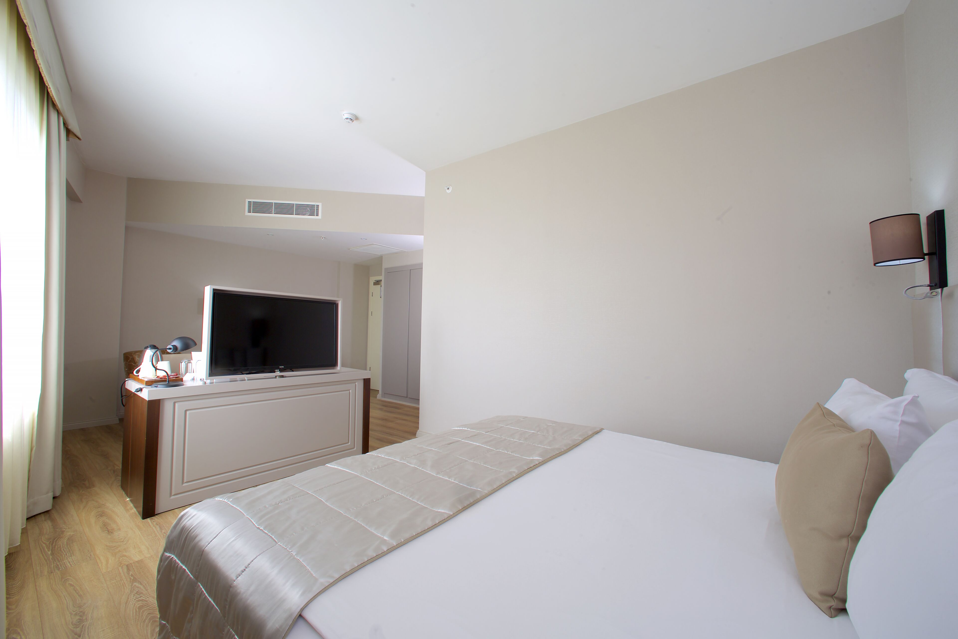 A hotel room with a large bed, a flat-screen TV, a desk with a lamp, and a wall-mounted TV.