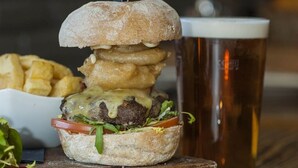 Hamburguesa con pan, queso, lechuga y tomate, acompañada de papas fritas y una cerveza sobre una mesa de madera.
