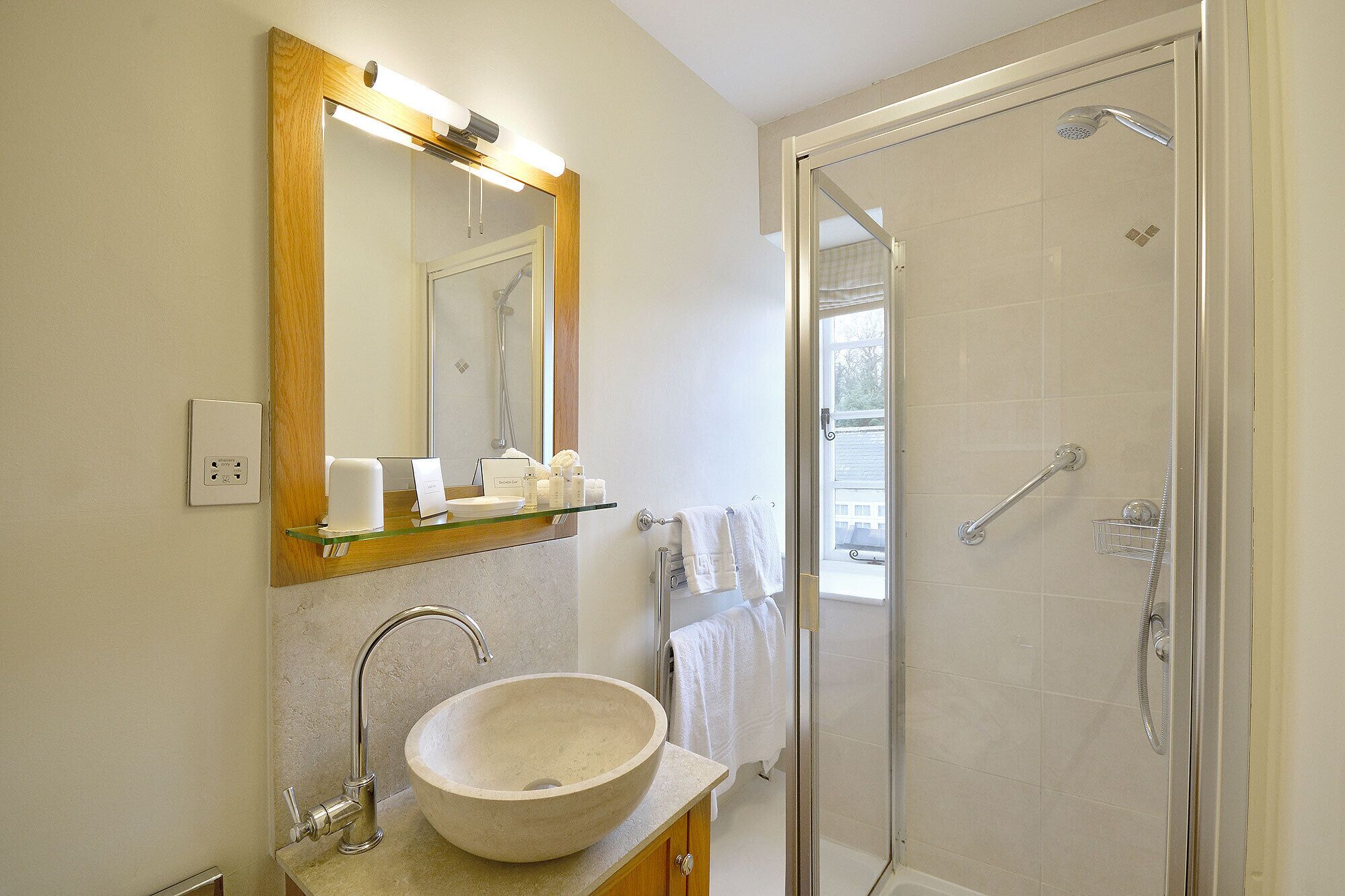 A bathroom with a large mirror, a vessel sink, a glass shelf with toiletries, a towel rack, and a walk-in shower.