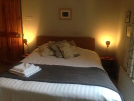 Una cama bien hecha con una manta gris y almohadas blancas, dos lámparas de noche, un cuadro enmarcado en la pared y una puerta de madera.