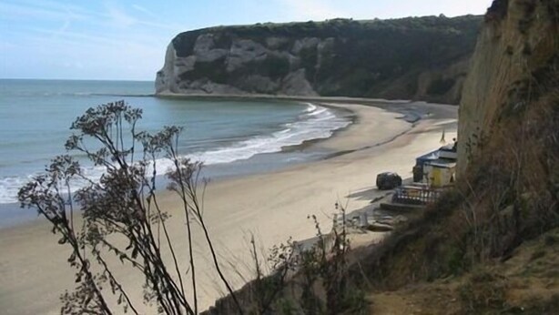 Paisaje costero con una playa de arena, un acantilado y un pequeño edificio cerca de la orilla.