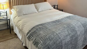 Ein ordentlich bezogenes Bett mit weißer Decke und grauer Wolldecke, ein Holz-Kopfteil, eine Wandkarte und eine Lampe auf einem Nachttisch.