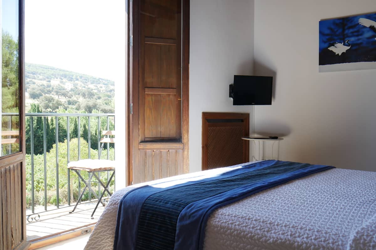 A bedroom with a bed, a painting, a television, and a balcony with a chair.