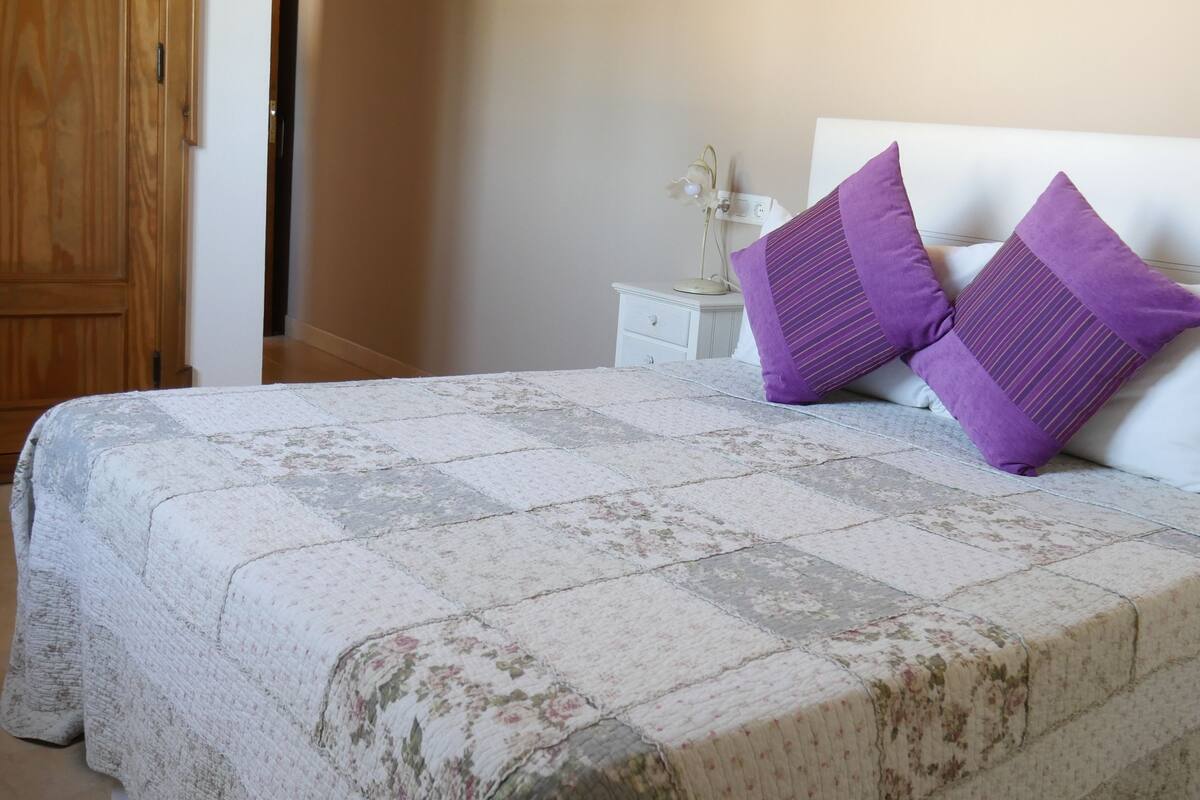 A bed with a floral-patterned bedspread, two purple pillows, and a white headboard.
