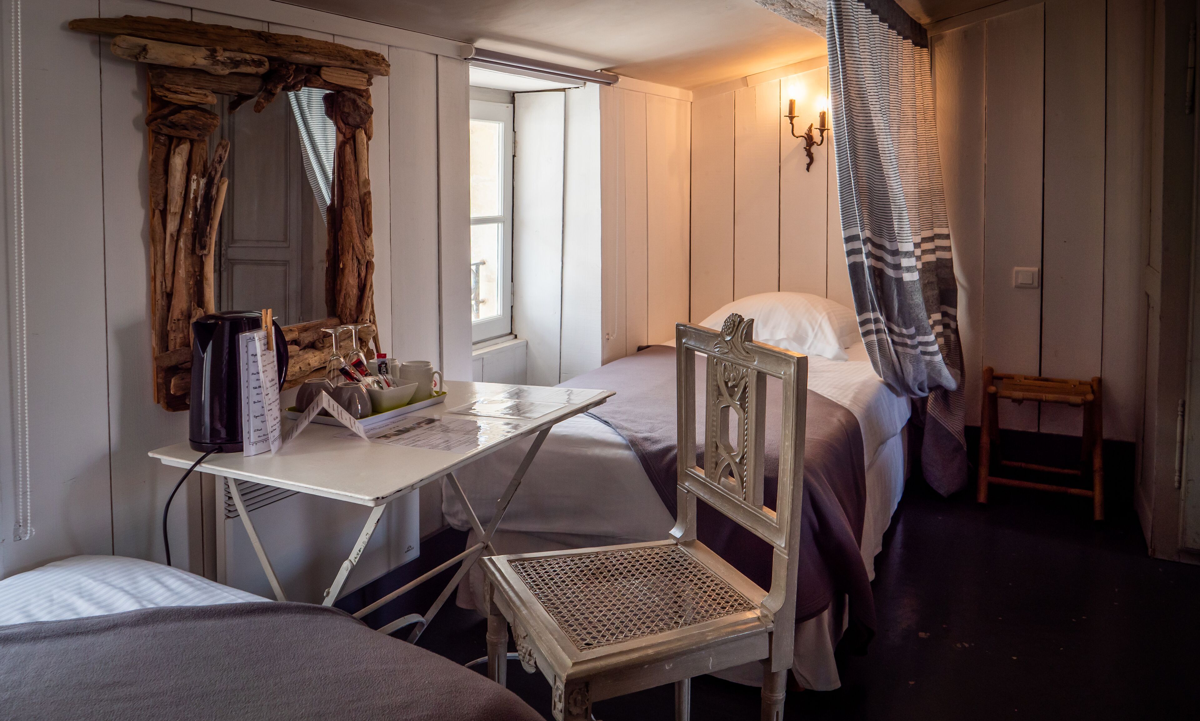 A room with a bed, a small table with a kettle, a chair, and a window.