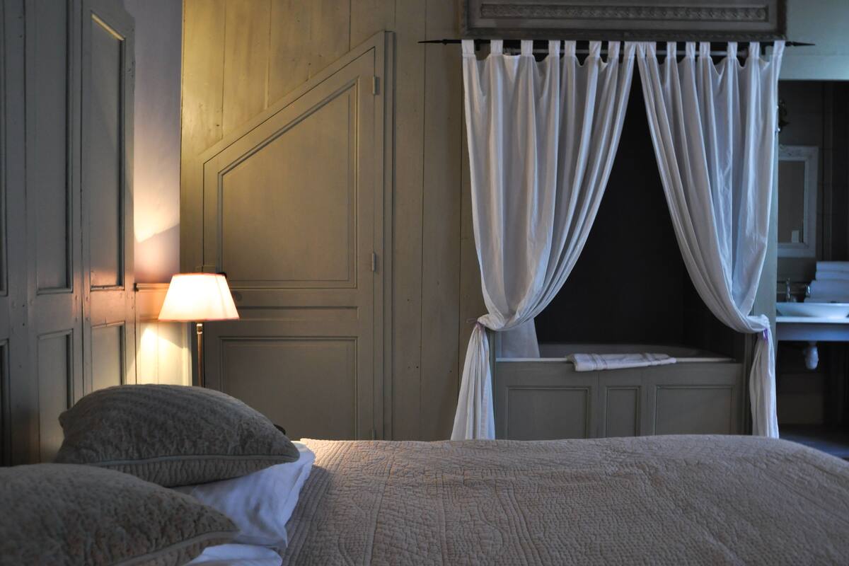 Ein Schlafzimmer mit Bett, Nachttisch mit Lampe, einem Fenster mit Vorhängen und einem im Hintergrund sichtbaren Badezimmer.
