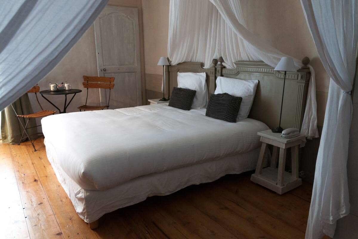 Ein großes Bett mit weißer Bettwäsche, ein hölzernes Kopfteil und zwei Nachttische mit Lampen. In der Ecke steht ein kleiner Tisch und zwei Stühle.