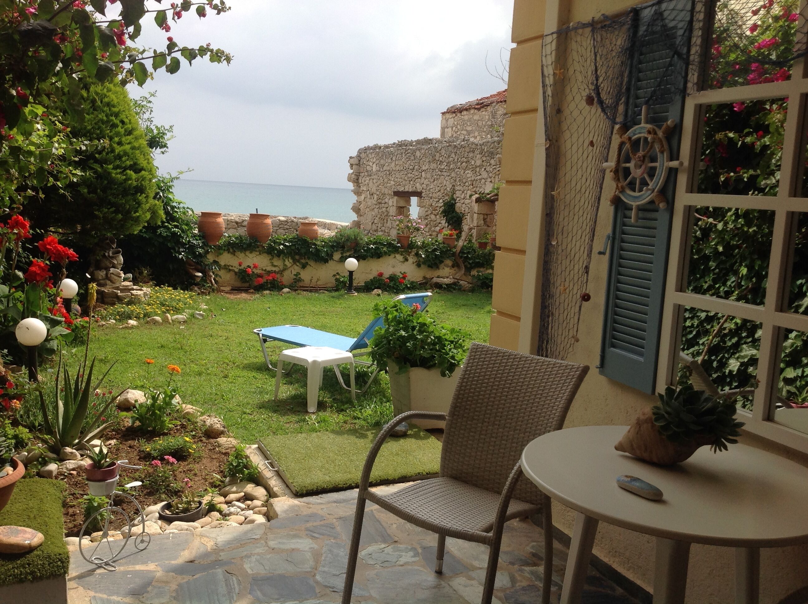 A patio with a table and chairs, a view of the sea, and a garden with potted plants and flowers.