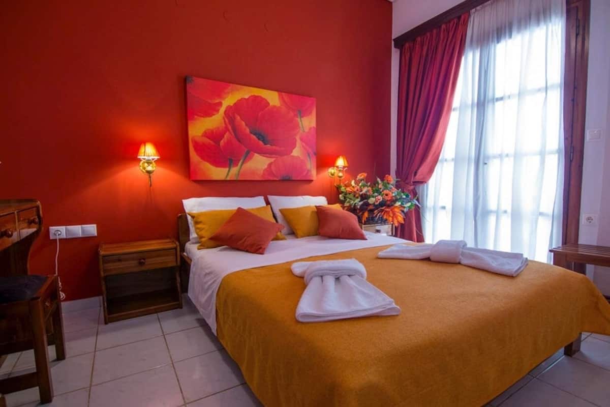 Habitación de hotel con una cama grande, paredes rojas, un cuadro de flores rojas, dos lámparas de noche, una mesita de noche de madera y un escritorio con una silla.