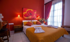 A hotel room with a large bed, red walls, a painting of red flowers, two bedside lamps, a wooden nightstand, and a desk with a chair.