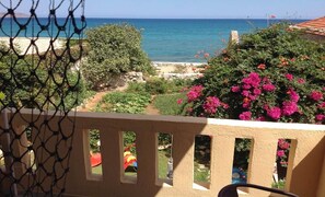 A balcony with a view of the sea, a garden with pink flowers, and a sandy beach.