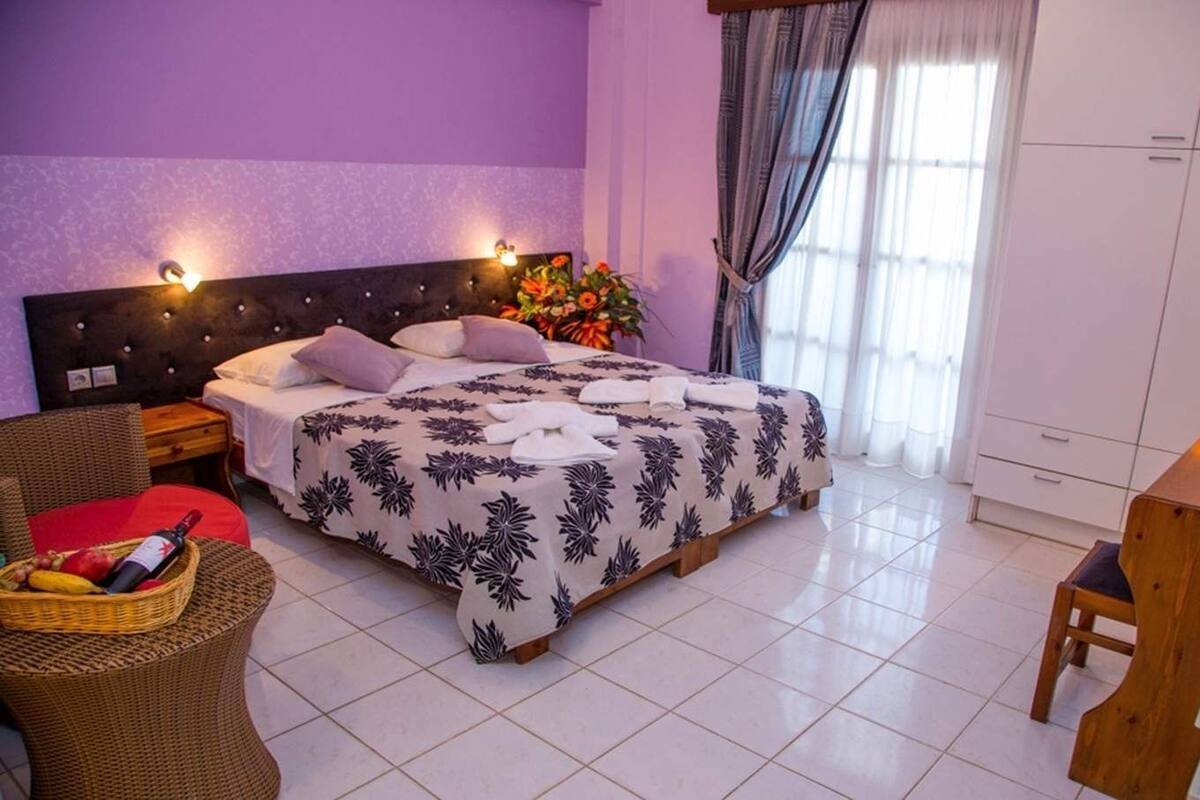 Un dormitorio con una cama, una silla de mimbre, una mesita con un florero y un armario.
