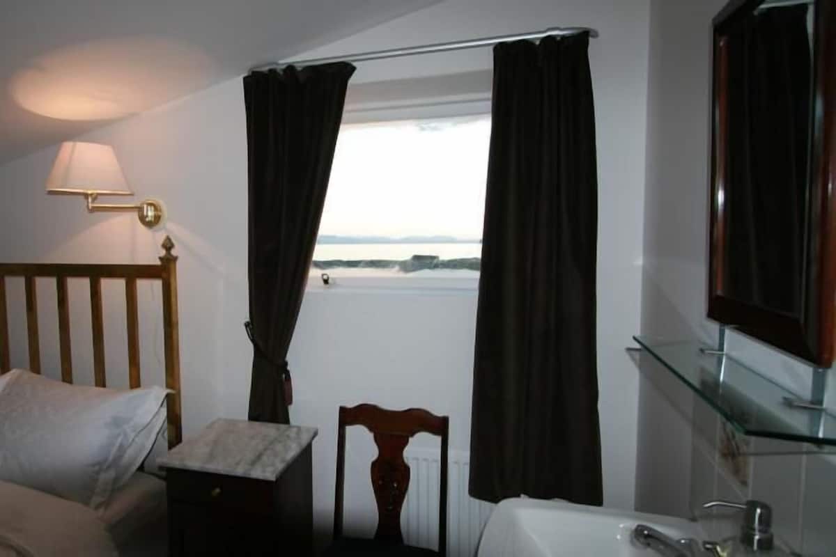 A bedroom with a bed, a nightstand, a lamp, a mirror, and a view of the sea through a window with curtains.
