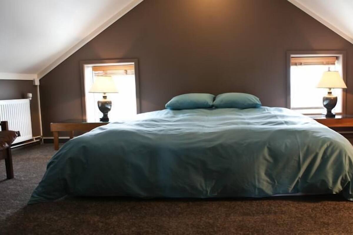 A bedroom with a large bed, two bedside lamps, a bench, and a radiator.